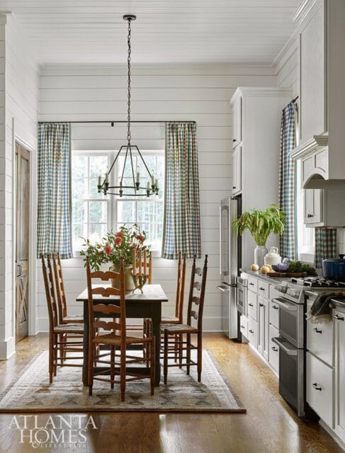 Farmhouse kitchen with white shiplap walls, a dark wood dining table with six chairs, plaid curtains, and white cabinetry with stainless steel appliances.