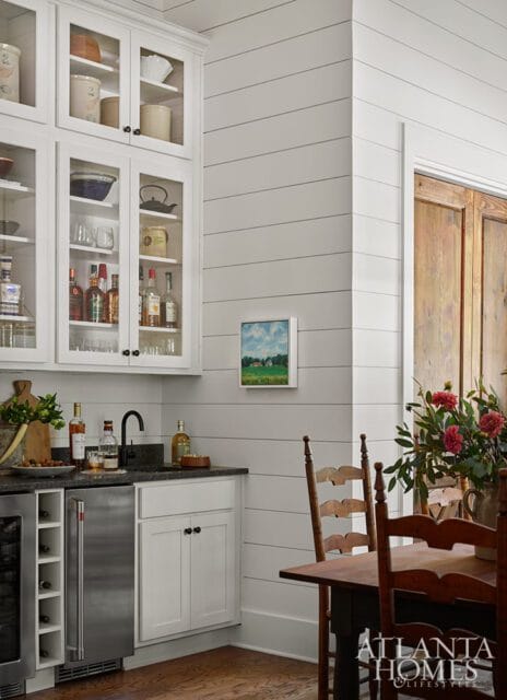 White shiplap walls in a kitchen area with glass-front cabinets filled with dishes and bottles, a wine fridge, and a small painting of a house on the wall.
