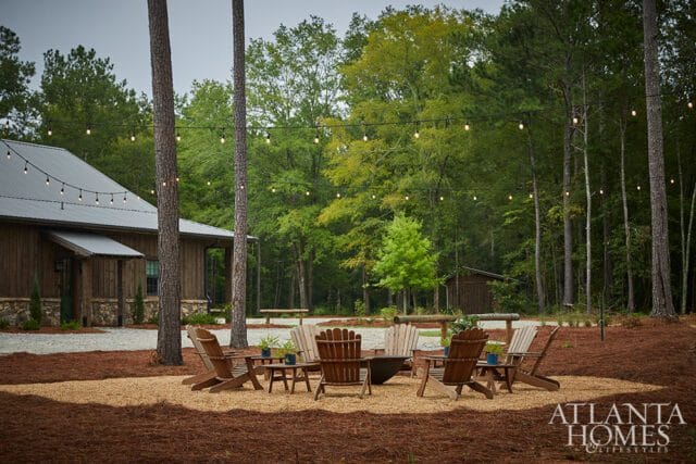 Rustic wooden building with metal roof, surrounded by pine trees and a gravel patio with Adirondack chairs and a fire pit. String lights hang overhead.