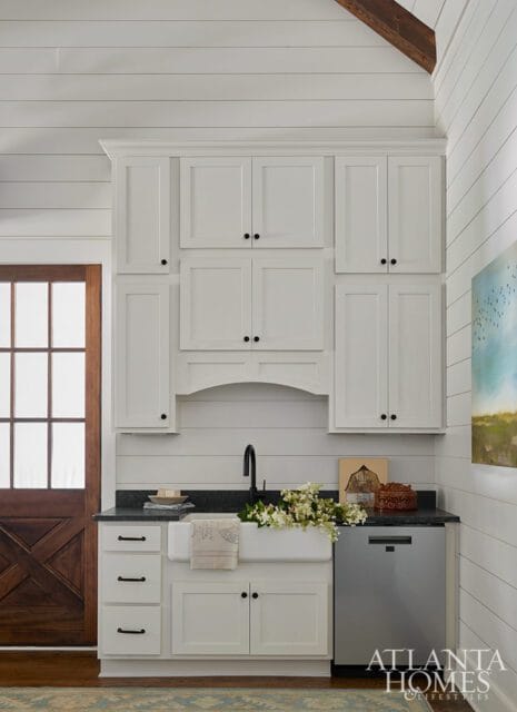 White kitchen with shiplap walls, white shaker cabinets, a farmhouse sink, black faucet, and a stainless steel dishwasher.