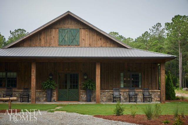 Wooden barn-style house with stone foundation, metal roof, green double doors, and rocking chairs on the porch.