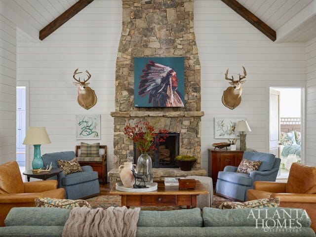 Living room with stone fireplace, mounted deer heads, leather armchairs, blue upholstered chairs, and a sectional sofa.