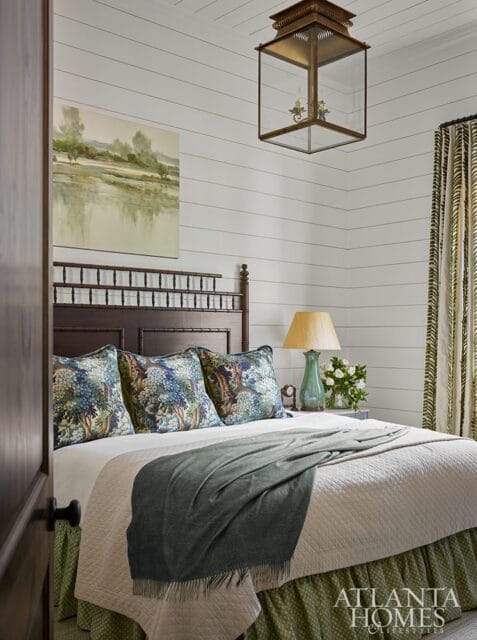 Bedroom with shiplap walls, a dark wood headboard with bamboo accents, and a bed dressed in a quilted coverlet, green throw, and botanical print pillows. A landscape painting hangs above the bed.
