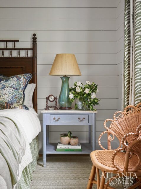 Bedroom with shiplap walls, a dark wood bed frame, a light blue nightstand with a green lamp and white flowers, and a wicker chair.