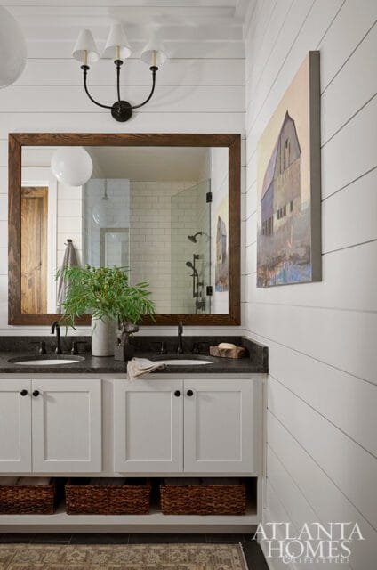 White shiplap bathroom with a double vanity featuring dark granite countertops, white shaker cabinets, and woven storage baskets below. A large wooden framed mirror reflects a subway tiled shower.