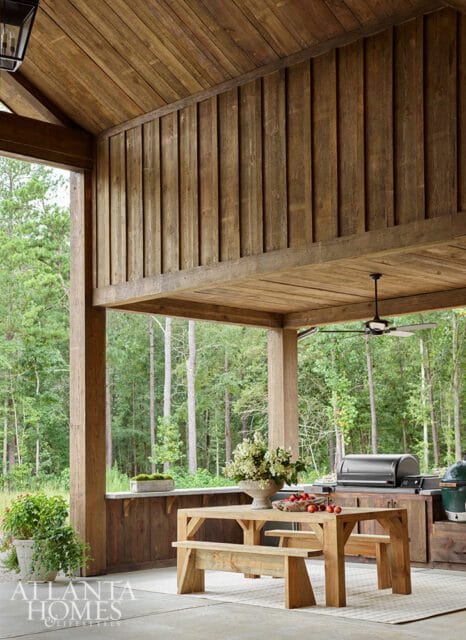 Covered porch with wood-paneled ceiling and walls, featuring a wooden picnic table, grill, and a lush forest view.