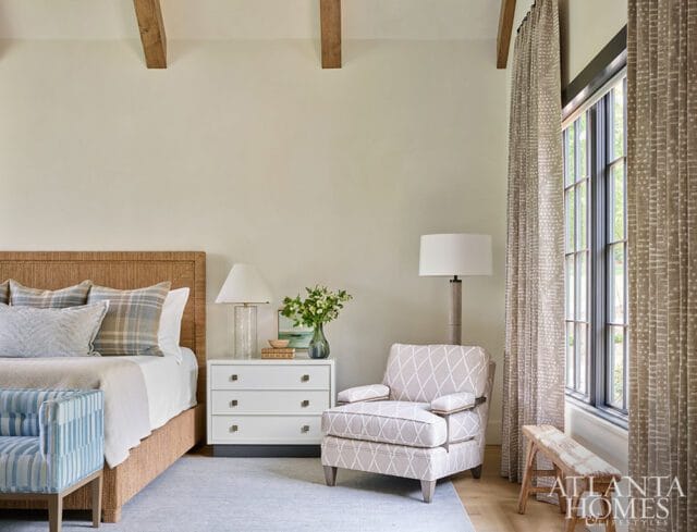 Bedroom with a woven headboard, plaid pillows, white nightstand, patterned armchair, and a large window with patterned curtains.
