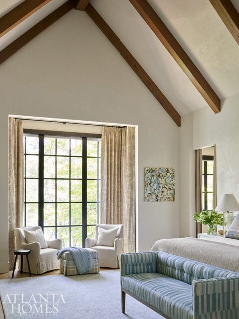 Bedroom with vaulted ceiling featuring exposed wooden beams, a large window with a view of trees, two upholstered armchairs, an ottoman, a striped bench, and a bed with a textured headboard.
