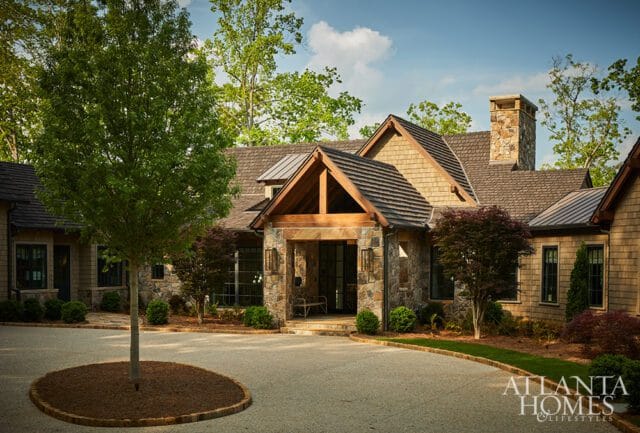 Stone and wood-shingled house with a covered entrance, stone chimney, and surrounding trees.