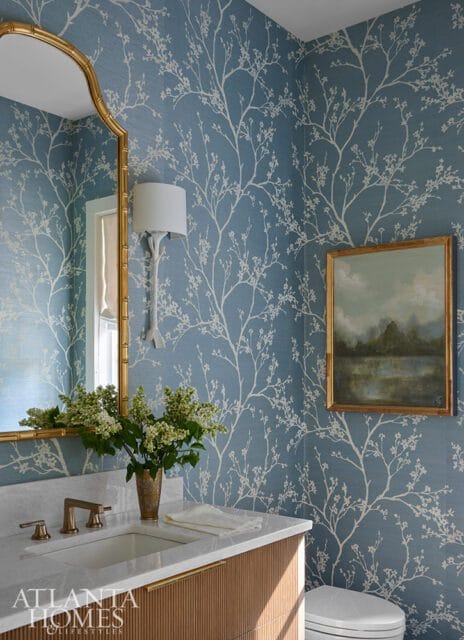 Powder room with blue floral wallpaper, marble vanity with brass fixtures, gold mirror, and a landscape painting.