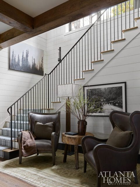 Interior staircase with shiplap walls, dark wood beams, black metal railing, and two leather wingback chairs with throw pillows and blankets.