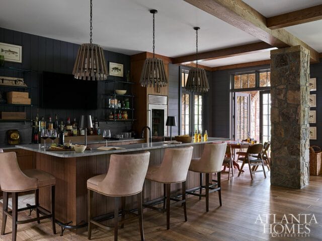 Bar area with gray stone countertop, dark wood cabinetry, and upholstered bar stools. Wooden pendant lights hang above. A stone pillar and dining area are visible in the background.