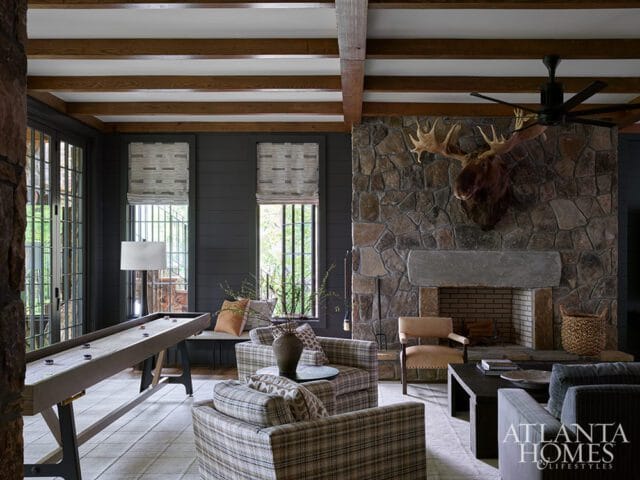Dark paneled living room with stone fireplace, mounted moose head, shuffleboard table, plaid armchairs, and Atlanta Homes & Lifestyles logo