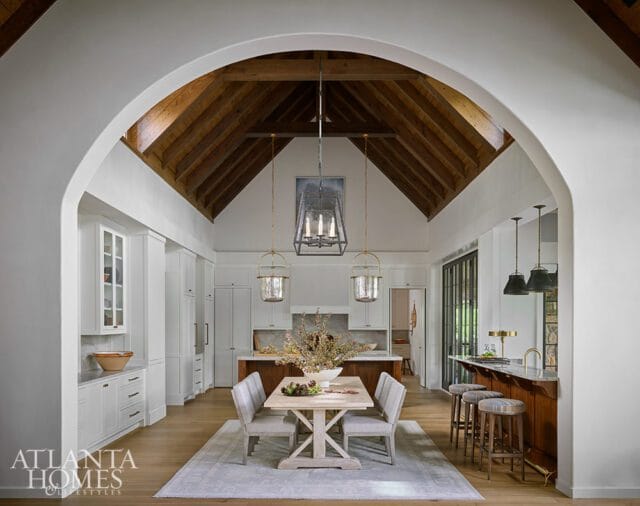 Vaulted wood-beamed ceiling above a dining area with a light wood table and upholstered chairs, leading to a white kitchen with marble countertops and a wood island with bar stools.