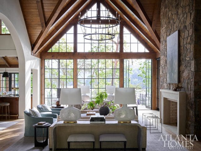 Vaulted living room with exposed wooden beams, stone fireplace, and large windows overlooking a wooded landscape.