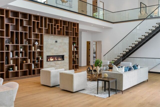 Modern living room with a large geometric wood bookshelf, stone fireplace with a fire, light gray sectional sofa with blue and white pillows, and a floating staircase with glass railings.