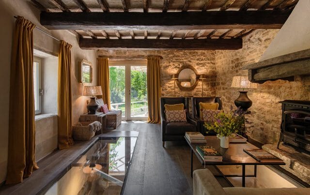 Living room with stone walls, exposed wooden beams, dark wood floors, mustard yellow curtains, two leather armchairs with patterned pillows, and a fireplace.