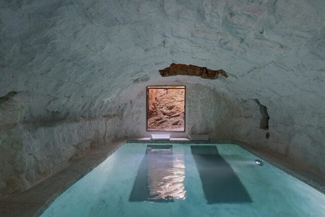 Indoor pool with rough, white-washed cave-like walls and ceiling, and a rectangular window looking out to a rocky exterior.