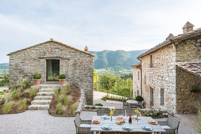 Stone buildings with tile roofs and a dining table set for a meal on a gravel patio, overlooking a lush green valley and mountains.
