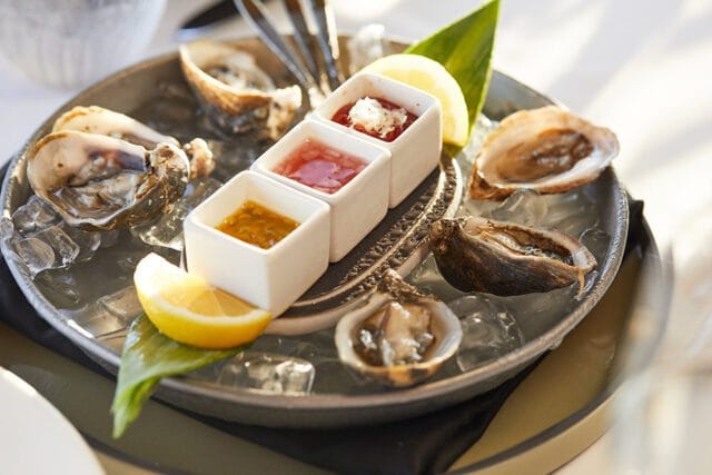 Plate of oysters on ice with three dipping sauces in white ramekins, garnished with lemon wedges and a green leaf.