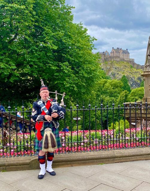 Man in traditional Scottish kilt and uniform playing bagpipes with Edinburgh Castle in the background.