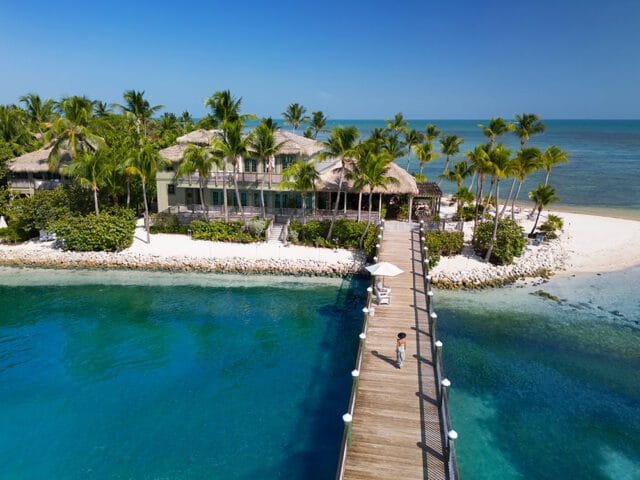 Tropical island resort with thatched-roof buildings, palm trees, white sand beaches, and a wooden pier extending into turquoise water.