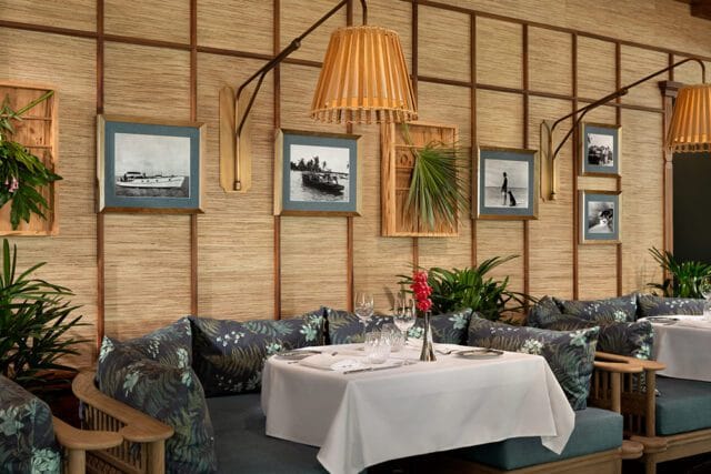 Dining room with bamboo-paneled walls, upholstered banquettes with tropical floral cushions, and tables set with white linens. Framed black and white photographs of boats and beach scenes adorn the walls.