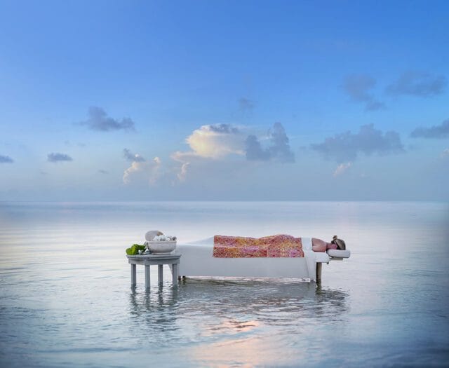Massage table set up in shallow ocean water under a blue sky with scattered clouds. A person lies on the table covered with a colorful blanket.