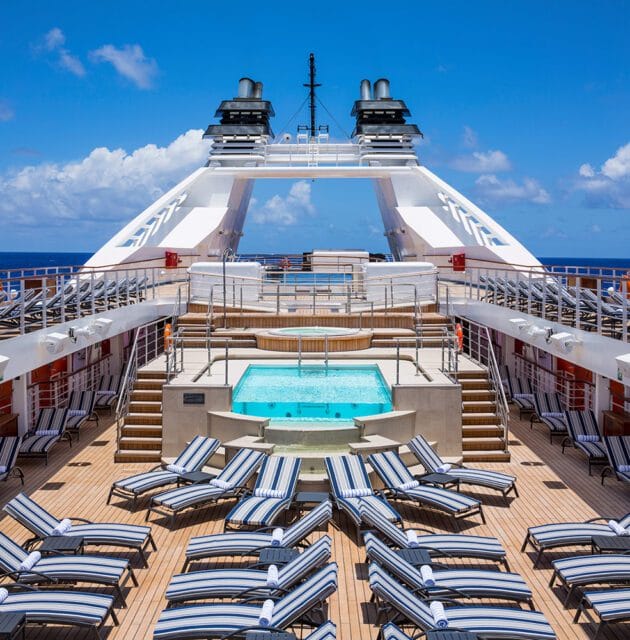 Deck of a cruise ship with lounge chairs surrounding a pool and hot tub, with the ocean and sky in the background.