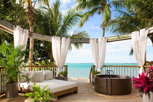 Wooden deck with a daybed, copper tub, and white curtains overlooking the ocean with palm trees