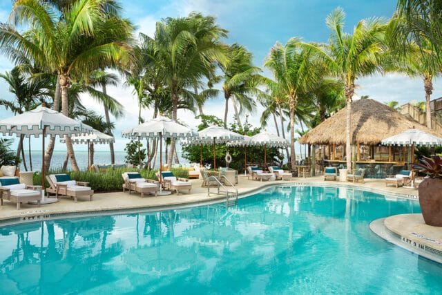 Resort pool area with lounge chairs, white umbrellas, and a thatched-roof bar surrounded by palm trees and overlooking the ocean.
