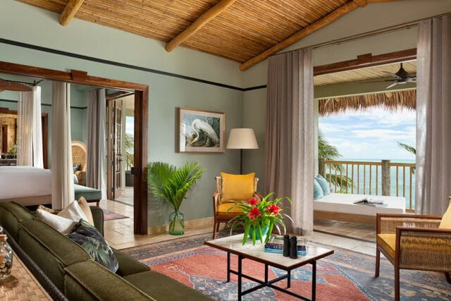 Living room with sage green walls, bamboo ceiling, and a large window overlooking the ocean. Features a green sectional sofa, patterned rug, and wicker furniture.