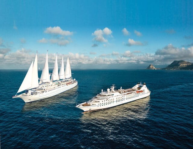 Two white cruise ships sailing on a deep blue ocean under a partly cloudy sky. One ship has five tall white sails.