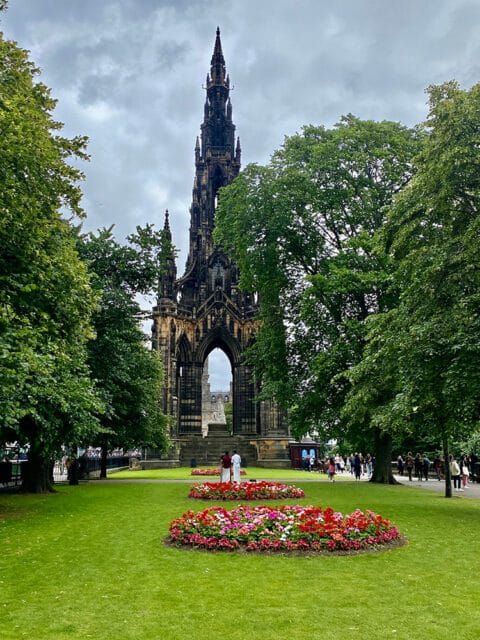 Scott Monument, a tall Gothic Revival spire, surrounded by lush green trees and manicured lawns with colorful flower beds in the foreground.