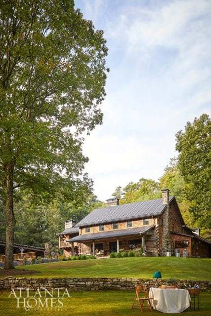Log and stone house with a metal roof, covered porch, and multiple chimneys, surrounded by trees and a manicured lawn.
