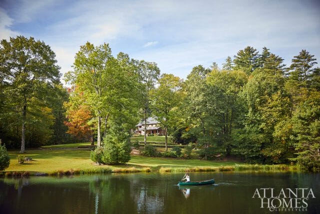 A person in a canoe paddles across a dark lake, with a large house nestled among trees on a grassy hill in the background under a partly cloudy sky.
