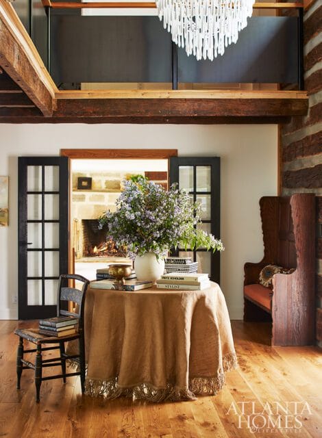 Round table with tan fringed tablecloth, books, and floral arrangement. French doors open to