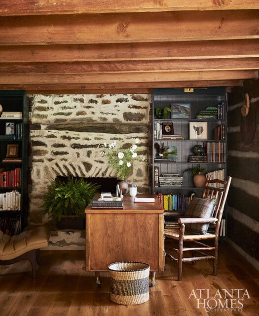 Wood desk with books and flowers in front of stone fireplace, flanked by bookshelves.
