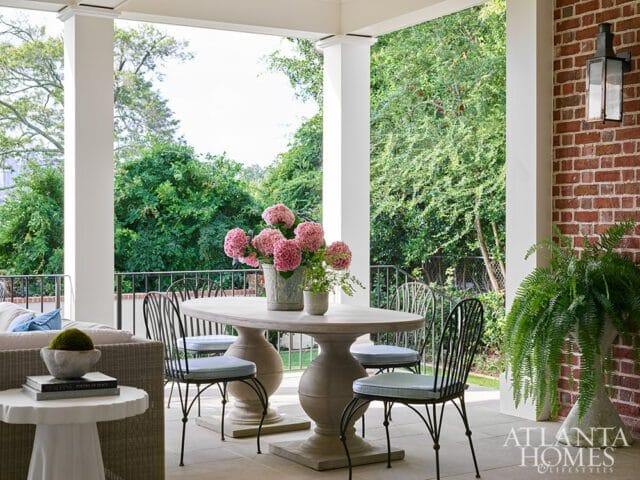 Covered patio with stone table and chairs, pink hydrangeas, and lush green trees in the background.