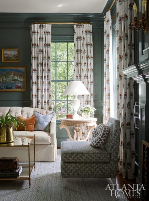 Living room with dark green walls, cream sofa with orange and blue pillows, patterned curtains, and a small armchair with patterned pillow.