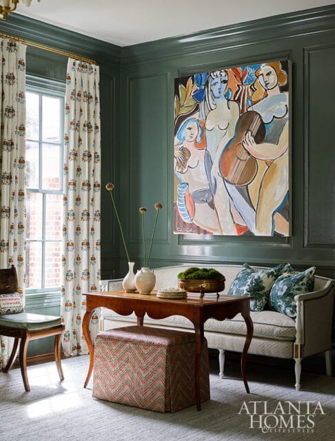 Dark green paneled walls with a large abstract painting of figures and a guitar above a sofa. A wooden table with vases and an ottoman sits in front of the sofa.