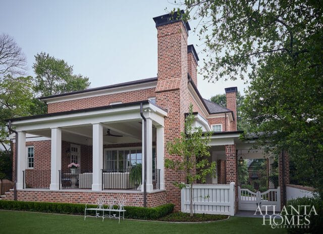 Red brick house with white columns supporting a covered porch, black metal railings, and two chimneys. A white picket fence and gate lead to the entrance.