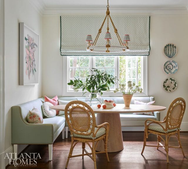 Light blue banquette seating with decorative pillows surrounds an oval wood dining table with two rattan chairs. A patterned roman shade covers a large window.