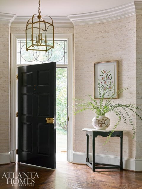 Entryway with a black paneled door, brass lantern, textured wallpaper, and a small console table with a vase of greenery.