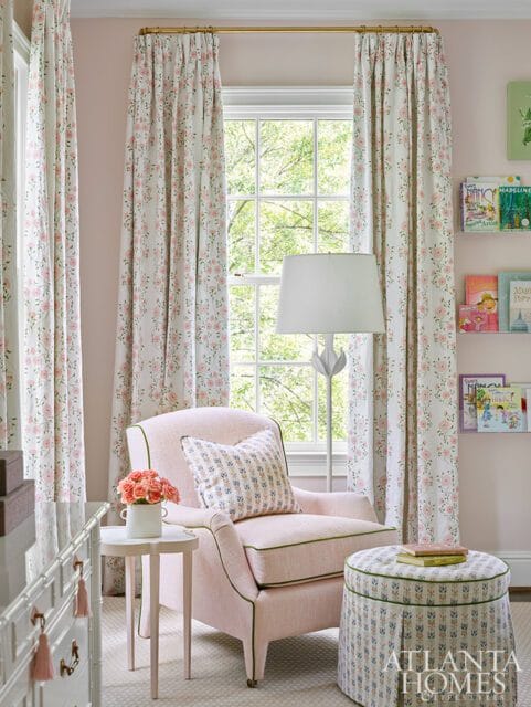 Pink armchair with floral patterned curtains, a small side table with roses, and a bookshelf filled with children's books.