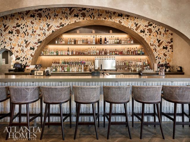 Bar area with a terrazzo backsplash, wooden arch, and shelves stocked with bottles. Woven bar stools with dark wood legs are in the foreground.