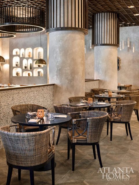 Restaurant dining room with round tables and woven chairs, textured walls, and decorative niches with pottery.