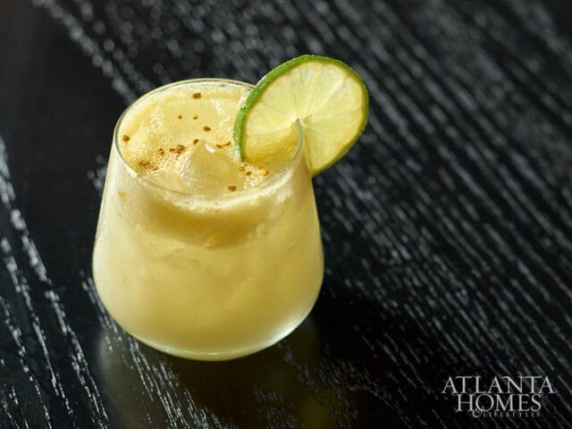 Yellow cocktail in a glass with ice, garnished with a lime wheel and sprinkled with spices, on a dark wooden surface.