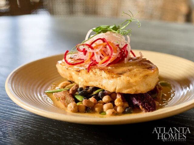 Seared fish fillet served over a medley of chickpeas, black beans, and green beans, topped with shredded red onion, carrot, and microgreens. Atlanta Homes & Lifestyles logo in the bottom right corner.
