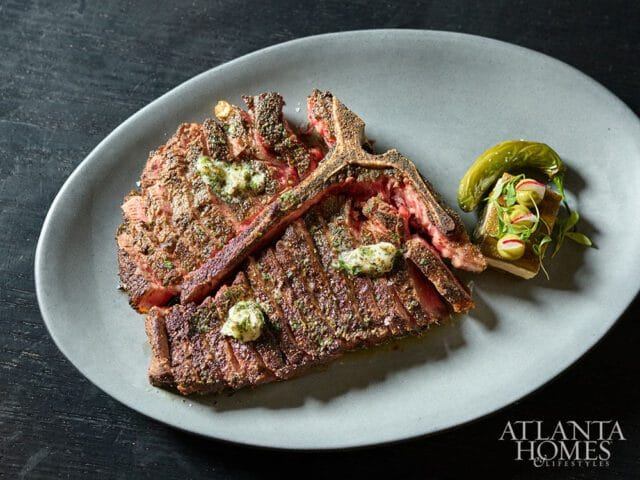 A thick-cut T-bone steak, sliced and topped with herb butter, served with a garnish of pickled peppers, olives, and radish on a grey plate.